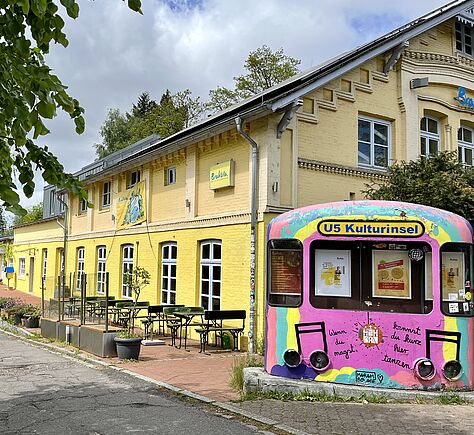 Vorn die Front einer U-Bahn, bunt bemalt, mit Plakaten in den Fenstern. Oben steht "Kulturinsel". Im Hintergrund das gelb gestrichene Gebäude des Brakula mit Stuck.