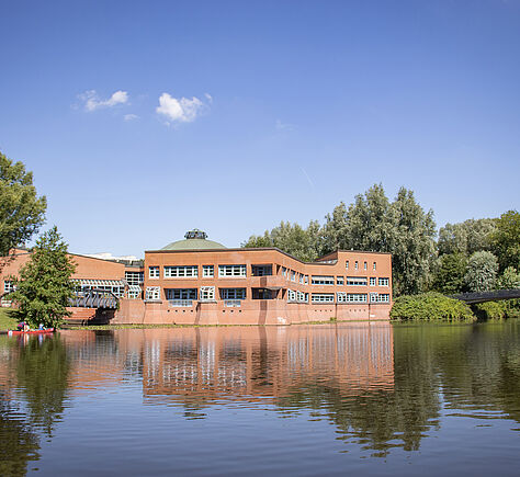 Im Vordergrund ein großer See, am hinteren Ufer ein flaches Backstein-Gebäude, roter Klinker. Links und rechts große Bäume