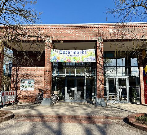 Ein Gebäude aus rotem Backstein mit großen Glasfenstern und einem Eingang in der Mitte. Über dem Eingang hängt ein Banner mit der Aufschrift „Ostermarkt“. Vor dem Gebäude stehen Fahrräder und Bäume werfen Schatten auf den Boden.