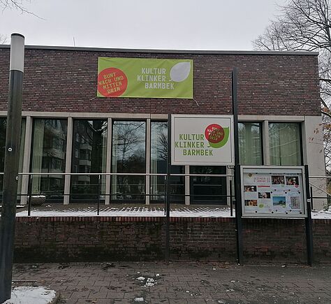 An der verklinkerten Gebäudefront ist ein grünes Banner angebracht mit dem Text Kultur Klinker Barmbek und einem weißen Blatt rechts daneben. Links davon ein roter Kreis mit dem Text: Bunt Wach Und Mitten Drin (alles in Großbuchstaben). Vor dem Gebäude ein weißes Schild mit dem selben Text