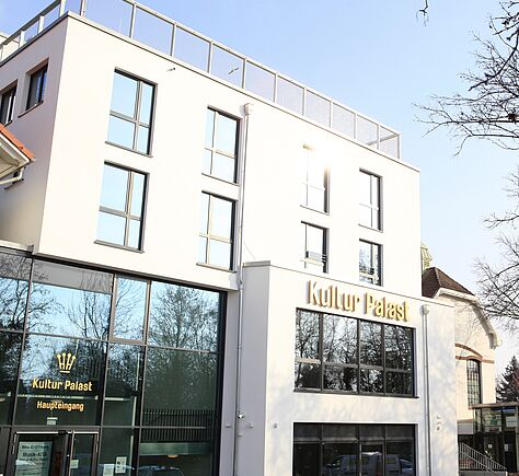 Moderner weißer Gebäudekomplex mit großen Fenstern und dem großen gelbgoldenen Schriftzug 'Kultur Palast' mittig rechts. Links unten der verglaste Eingangsbereich, ebenfalls beschriftet mit 'Kultur Palast' darüber das Kronen Logo und darunter kleiner 'Haupteingang'.