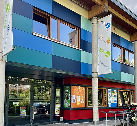 Teilansicht der Fassade des Stadtteilzentrums mit einer quadratischen Verkleidung in verschiedenen Blautönen. Zwei vertikale Banner an den Säulen beim Eingang. Darauf steht Kultur A. Beim Eingang sind mehrere Fahrräder an einem Fahrradständer abgestellt.