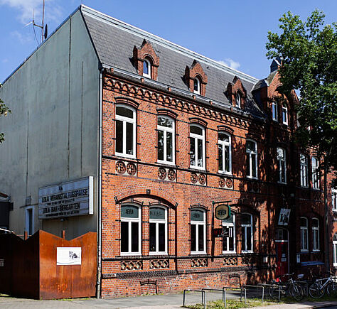 Ein altes rotes Backsteingebäude am Straßenrand mit vielen weißgerahmten, leicht bogenförmigen Fenstern. Rechts davor ein großer Baum. Der Eingang zur LOLA befindet sich an der linken Seite in einem kleinen Anbau.