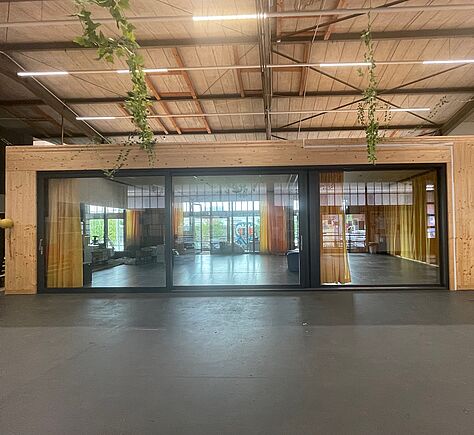 In eine große Halle ist der Tanzsaal hinein gebaut. Sein Rahmen und die Decke der Halle sind aus Holz. Die Front des Saales bildet eine Glas-Schiebetür. die fast über die gesamte Breit geht. An der Hallendecke hängen Girlanden aus grünen (Plastik-) Blättern
