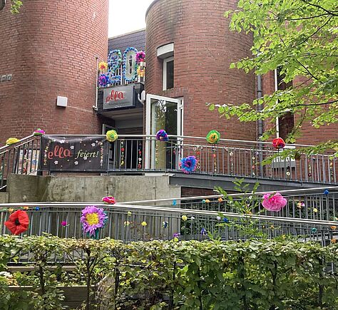 Der Eingang zu einem Backsteingebäude mit zwei runden, zylindrischen Gebäudeteilen und einer Rampe mit Metallgeländer, die mit bunten Papierblumen dekoriert ist. Über dem Eingang hängt eine große „20“ aus blauem Material mit bunten Deko-Elementen daneben. Unter der „20“ ist ein Schild mit dem Wort „ella“ in roter Schreibschrift; darunter steht in kleiner Schrift „Kulturhaus Langerhagen“. Am Geländer links hängt ein schwarzes Banner mit der Aufschrift „ella feiert!“ und kleinen Blumenmotiven.