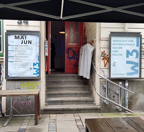Hauseingang mit einigen Stufen führt zu einer roten Tür. Links und rechts des Eingangs hängen zwei große, gerahmte Informationstafeln. Die linke Tafel zeigt einen Veranstaltungsplan, die rechte trägt die Aufschrift 'Werkstatt für internationale Kultur und Politik' sowie das Logo 'W3'.
