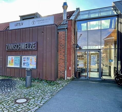 Rechts der gläserne Eingangsbereich der Zinnschmelze. Davor ein ebenmäßig gepflasterter Weg. Links angrenzend an den Glasbereich besteht das Gebäude aus roten Backsteinmauern, daran eine rostfarbene Metallverkleidung mit der Aufschrift "Zinnschmelze". Darunter bunte Plakate, darüber 2 weiße Schilder mit der Aufschrift "Lüttliv" und "Theaterdeck Hamburg". Vor diesem Teil des Gebäudes ist eine Fläche mit Kopfsteinpflaster und einem Poller.