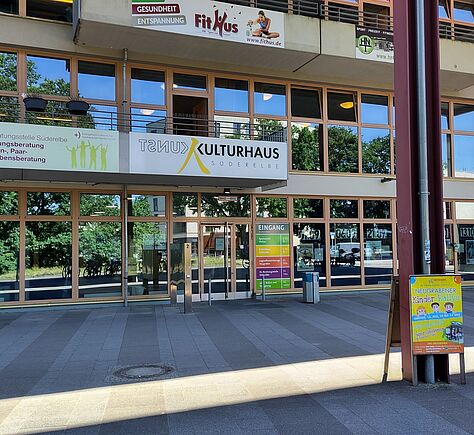 Über dem Eingang ein großes Schild mit dem Namen des Kulturhauses. Links daneben ein weiteres Schild, das auf die "Ev. Beratungsstelle Süderelbe", die Erziehungsberatung für Familien, Paare und Lebensberatung. Im oberen Bereich des Gebäudes ein weiteres Schild: von "Fit Hus". Vor dem Eingang steht ein Plakatständer, der für eine "Neugrabener Kinder Rallye" wirbt. Das Gebäude hat viele Fenster, in denen sich die Bäume vor blauem Himmel und Fahrräder vor dem Gebäude spiegeln.