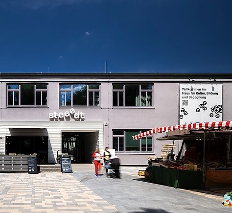 Ein modernes 2-stöckiges Gebäude mit großen Fenstern. Rechts eine große weiße Tafel auf der steht: Willkommen im Haus für Kultur, Bildung und Begegnung. Links neben dem Gebäudeeingang ein Café mit Außenterrassen, darüber der Name: "streedt"