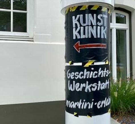 KUNSTKLINIK Eine Litfasssäule mit einem Plakat auf dem steht: KUNSTKLINIK (darunter ein roter Pfeil, der nach links zeigt), Geschichtswerkstatt, martini-erleben. Dahinter eine weiße Hausfassade mit weiß umrahmtem Fenster und einer Glastür.