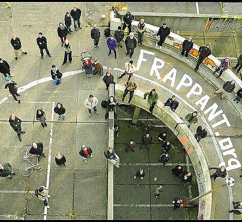 Blick von sehr weit oben: Viele Menschen stehen im Halbkreis. Schriftzug frappant.org ist auf dem Boden aufgemalt