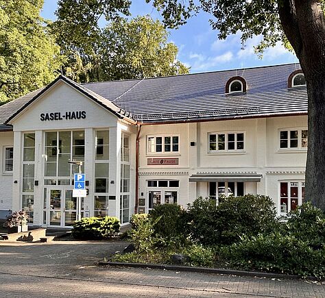 Außenansicht mit Eingang, das Haus hat eine weiße Fassade, 2 Stockwerke und viele Fenster. Das Dach ist mit grauen Ziegeln gedeckt. Über dem verglasten Eingang ein Schild 'SASEL-HAUS'. Ein belaubter großer Baum rechts im Bild. Rollstuhlparkplatz auf dem gepflasterten Vorplatz.