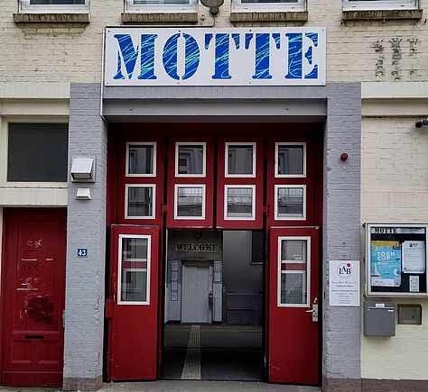 Das Bild zeigt den Eingang zum Innenhof der "MOTTE" (steht in großen blauen Buchstaben auf einem weißen Schild über der Tür). Eine sehr große rote Tür mit vielen weiß gerahmten Fenstern. Links neben der Tür ist eine kleine rote Tür, rechts ein Schaukasten mit verschiedenen Aushängen und Plakaten. Über dem Eingang sind drei Fenster mit dem Logo "LAB" (Lange Aktiv Bleiben e.V.). Die Hausnummer 43 ist links neben der Tür angebracht. Auf der gegenüberliegenden Seite des Innenhofes ist über dem Fahrstuhl ein Schild mit der Aufschrift "WELCOME" zu sehen.