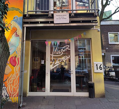 Der ebenerdige Eingangsbereich eines gelben, 2-stöckiges Hauses. Auf der linken Seite wächst sehr viel Efeu an der Hauswand und verdeckt halb ein Wandbild, auf dem essen zu erkennen ist. Über der eingangstür ist ein Balkon, an dem ein Schild hängt mit der Aufschrift: "Hamburg-Mitte. St. georg. Ein Ort der Vielfalt."