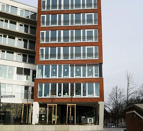 Mehrstöckiges rechteckiges Gebäude mit rotem Backstein, Flachdach und vielen großen Fenstern. Links schließt sich schräg ein weißes Haus in ähnlichem Stil an. Im Vordergrund junge Bäume und Gehweg.