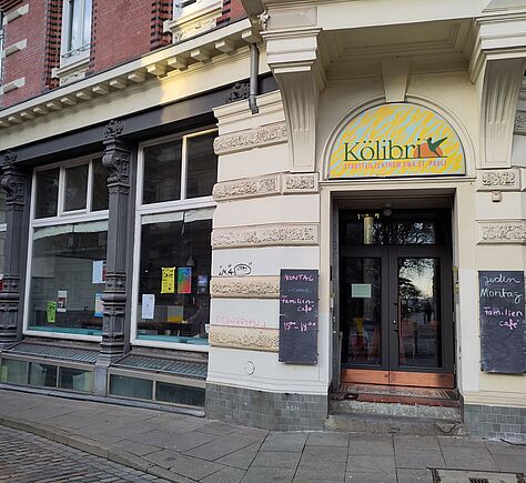 Ein Altbaugebäude über Eck mit heller Fassade mit Verzierungen, links mit dunklen Säulen und sehr großen, botentiefen Fenstern. Gelbes Logo über der verglasten Tür und zwei schwarzen Tafeln mit handgeschriebenen Öffnungszeiten rechts und links neben dem Eingang.