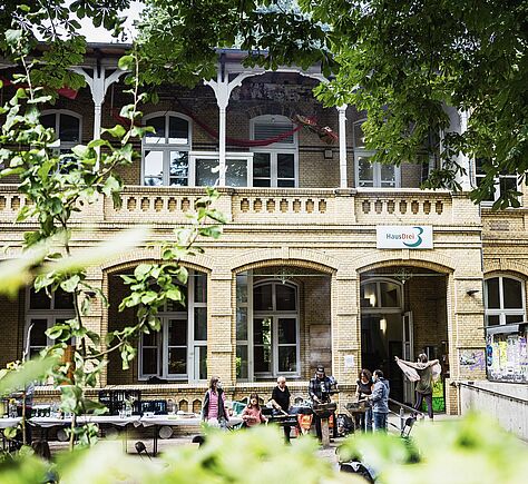 Ein Altbau, zweistöckig mit Arkaden, umgeben von grünen Pflanzen, auf dem Hof davor sind Tische aufgebaut, an denen Menschen sitzen