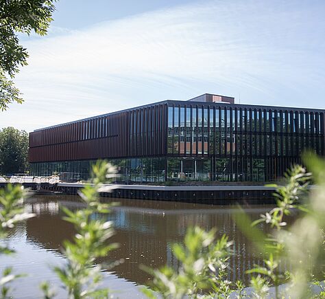 Das Gebäude von außen. Ein moderner Bau, viel Glas und mit Flachdach. Das Gebäude von Wasser umgeben, im Vordergrund sind unscharf Pflanzen.